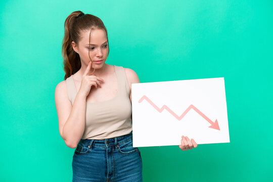 Young caucasian woman isolated on green background holding a sign with a decreasing statistics arrow symbol and thinking