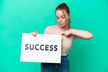 Young caucasian woman isolated on green background holding a placard with text SUCCESS and  pointing it