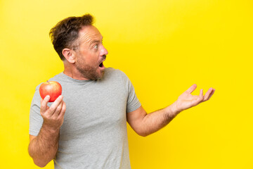 Middle age caucasian man with an apple isolated on yellow background with surprise facial expression
