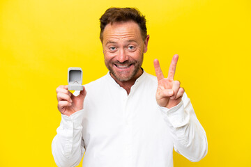 Middle age man holding a engagement ring isolated on yellow background smiling and showing victory sign