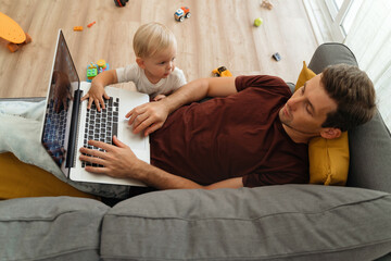 Naughty little baby boy bothering his father lying on couch with laptop in living-room, tapping keyboard with hand, disturbing working from home or resting dad. Toddler son asking for dad attention