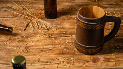 Oak mug with a handle and a glass brown bottle on a wooden table in a pub or bar. Handmade wooden tankard for beer or kvass. Brown mug with ornament, wheat grains and spikelet on a wooden surface.