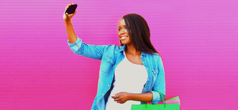 Portrait of happy smiling african woman with shopping bags taking selfie with smartphone on pink background
