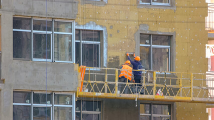 Workers are insulating the wall of a building. Construction site with working men on the elevator....