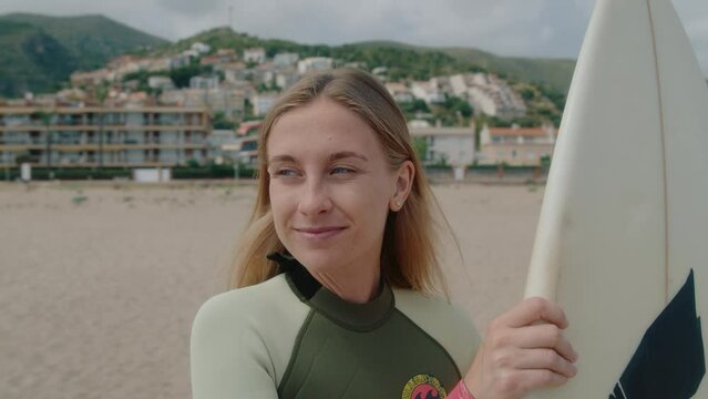 Portrait of young caucasian woman with surfboard, smile and look into camera, happy and motivated summer adventure. Surf camp in holiday resort. Surfing classes and surf vacation