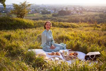 A girl in a long summer dress with short hair sitting on a white blanket with fruits and pastries and enjoying the moment. Concept of having picnic in a city park during summer holidays or weekends. 