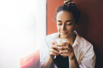 Young woman drinking coffee cappuccino sitting near window.