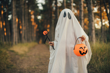 Girl wearing ghost costume holding pumpkin bucket with candies, standing in a forest.
