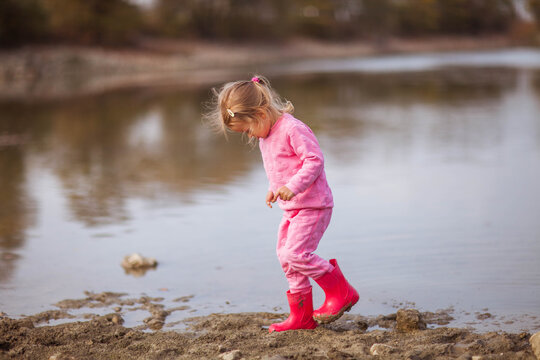 Girl in a pink suit and red rubber boots walking and throwing pebbles in lake. Nature autumn photo backgrounds