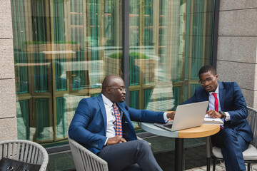 Two dark-skinned African American businessmen in suits and glasses with briefcases sit at a table in an outdoor city cafe.