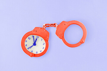 Vibrant orange handcuffs with a clock on isolated vivid purple background. Business abstract concept. Minimal flat lay. The idea of lack of time, deadline or trapped in time. Law and justice symbol.