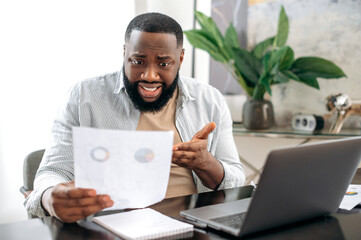 Confused puzzled african american business man, freelancer or company ceo, working with documents, sit at desk in modern office, working on a project uses laptop, studying financial graphs, stunned