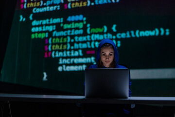 Hooded anonymous hacker woman by computer in the dark room at night, cyberwar concept.
