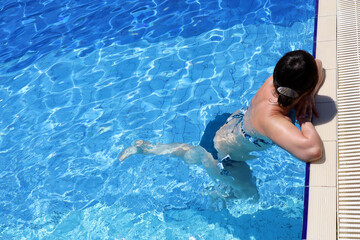Woman in swimsuit relax in transparent water leaned on edge of swimming pool, top view. Girl swimmer in summer, beach vacation and cooling in hot weather