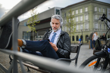 Fototapeta premium Businessman with bike sitting on bench, listening to music with feet up and resting. Commuting and alternative transport concept