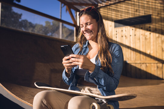 Image of happy caucasian woman with smartphone resting in skate park