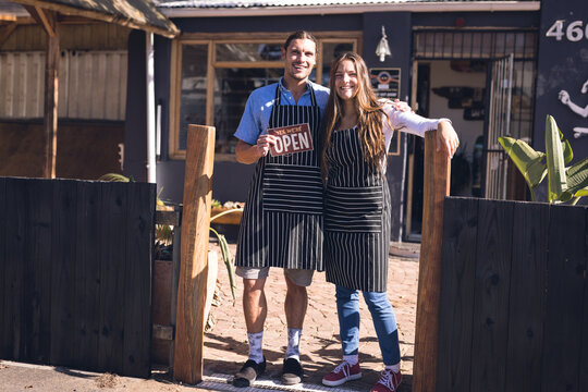 Image of happy caucasian woman and man in aprons and open sign in front of skate shop
