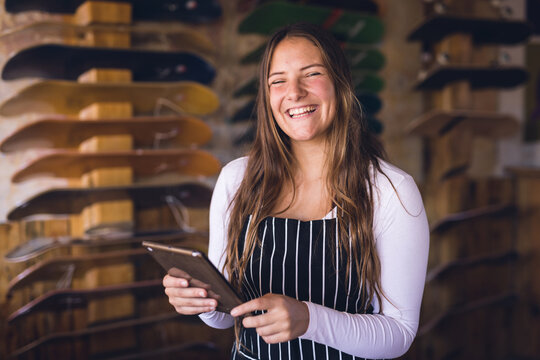 Image of happy caucasian woman in apron with tablet posing in skate shop