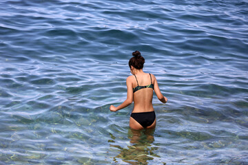 Girl in black swimsuit going to swim in sea water. Vacation on a pebble beach of summer resort