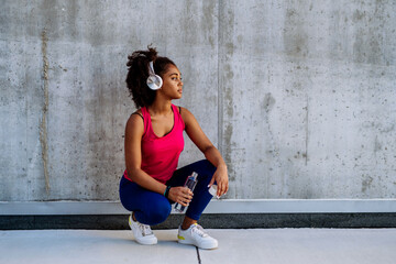 Young multiracial girl in sportswear resting after jogging in city, drinking water from sustainable bottle and listening music in headphones.