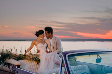 Young couple in love posing at sunset in nature on their wedding day near a red car