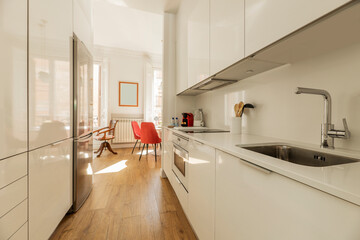 Fototapeta na wymiar Kitchen with gloss white wood cabinetry, white stone countertops, white built-in appliances and chestnut hardwood floors and access to a bright living room