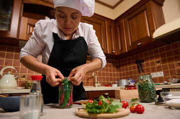 Beautiful stunning woman, housewife wearing white chef's cap and black kitchen apron inserting red and green hot chili in a sterilized glass jar, preparing fermented delicacies and marinated food