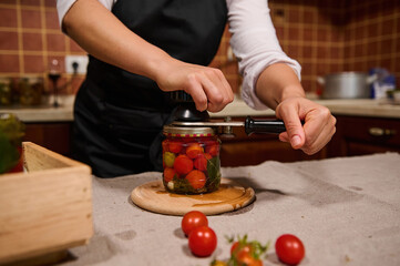 Details: housewife's hands using seamer, closes lids of a jar with fermented organic homegrown cherry tomatoes. Homemade canned food. Preserving fresh seasonal harvest for the winter. Mmarinating