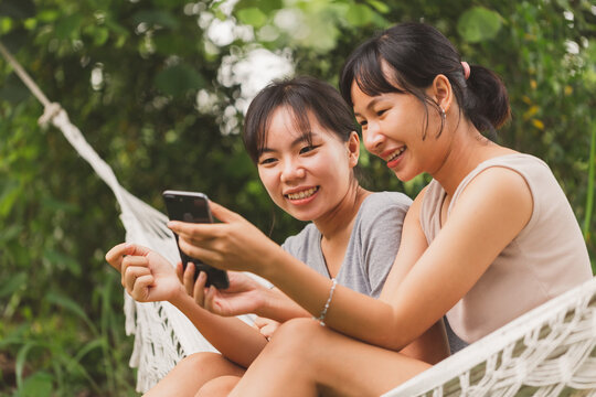 Young Asian teenager women sitting on hammock taking selfie outdoor rounded by green trees. Happy student friend enjoy vacation and taking picture together.