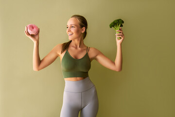  Adult sporty woman standing near a green background, holding a pink donut in one hand and broccoli...