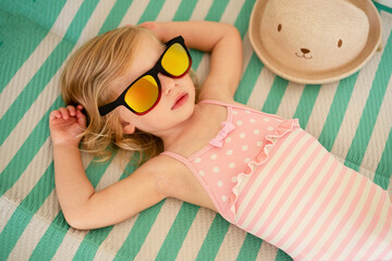 Blonde caucasian toddler, baby girl lying on striped towel on beach with sunglasses, swimsuit sunbathing,taking sun.Kid,white skin tan concept, child health care, copyspace.