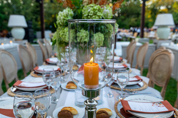 Beautiful decoration of a wedding banquet with a meal in the green garden in white colors. Very stylish setting with tropical flowers in a vase, glass candlestick set, napkins, plates, glasses cutlery