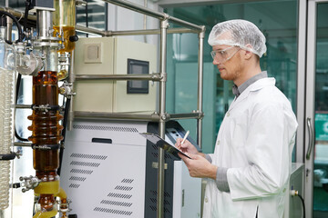 scientist writing on clipboard and looking cannabis extraction machines in testing lab