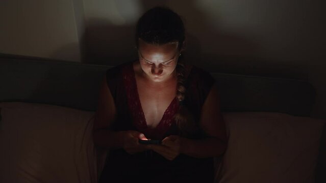 At night, the girl is sitting on the bed, holding the phone, laughing. View from above.