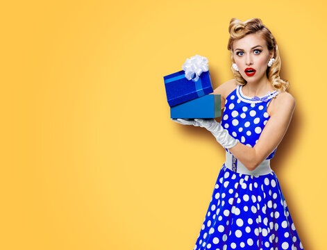 Image of beautiful woman dressed in pin up blue dress in polka dot white gloves opening gift box, isolated on yellow color background. Blond pinup model in retro fashion studio concept.
