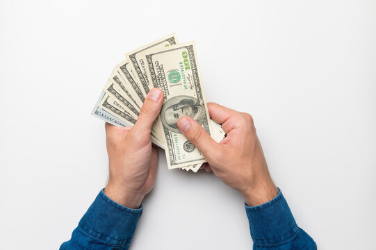 A first-person photograph showing male hands holding a fan of one hundred dollar bills against a white background. studio shot