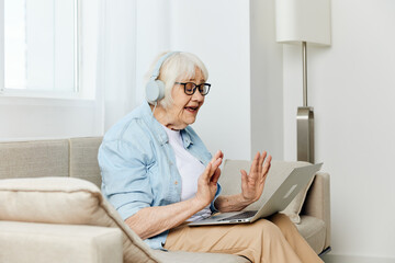 a happy, smiling elderly woman is sitting at home on a cozy sofa with headphones on her head, holding a laptop on her lap while working from home