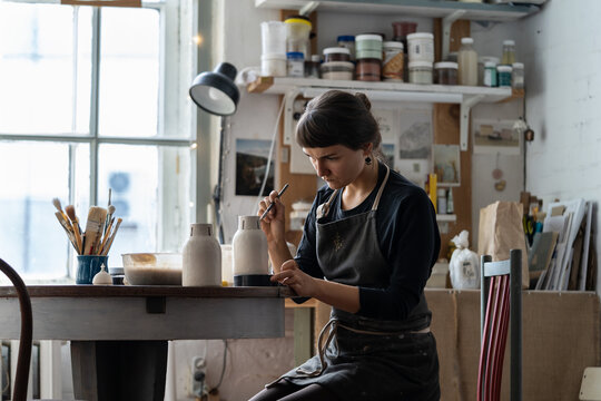 Young Female Artisan In Black Apron Works With Crockery Sitting At Round Table In Workshop Against Bright Window. Brunette Woman Enjoys Painting Handmade Ceramic Vase In Pottery Craft Studio