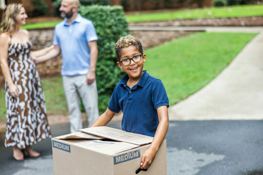 A little boy holding a moving box ready to move into his new house while parents talk in the background
