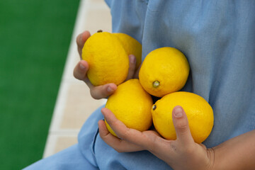 children's hands hold lemons, yellow lemons, blue denim sundress, green lawn
