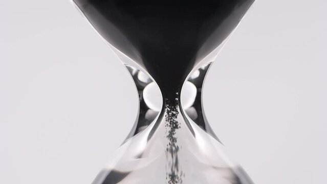 Grains of black sand and quartz are falling in a glass hourglass.