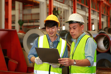 Two Asian male engineers inspect galvanized sheet metal coils before shipping using a laptop.