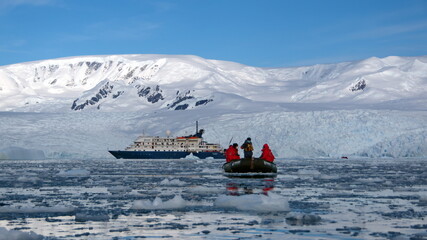 Zodiac inflatable boat navigating among icebergs at the base of a snow covered mountain, at Cierva Cove, Antarctica, with an expedition cruise ship in the background