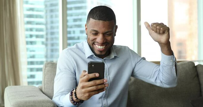 Cheerful African guy on couch at modern home read sms with fantastic commercial offer, get huge discounts, sell-out, celebrate great opportunity, feels overjoyed. Triumphant rejoicing success concept