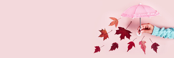 Woman's hand with pink umbrella and falling leaves on pink pastel background. Hello Autumn creative...