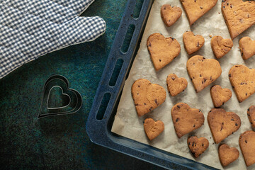 Heart shaped biscuits with chocolate drops on a baking tray near an oven glove and cake forms.