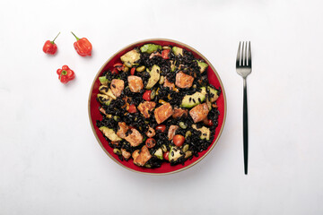A red bowl of black rice with baked salmon, avocado, tomatoes, chili peppers and edamame.