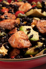 A red bowl of black rice with baked salmon, avocado, tomatoes, chili peppers and edamame.