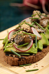 An avocado toast with mushrooms, radish, sesame seeds and chive on a wooden cutting board.