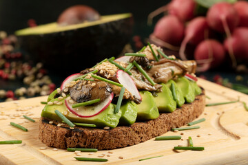 An avocado toast with mushrooms, radish, sesame seeds and chive on a wooden cutting board.